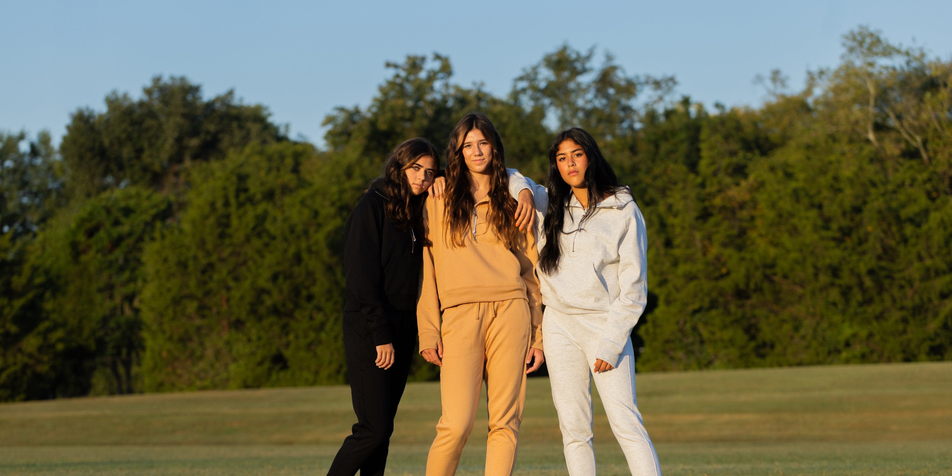 Three girls standing next to each other in a field outside while wearing XX-XY Fearless Fleece Zip and Jogger sets in the colors black, terra cotta, and white.